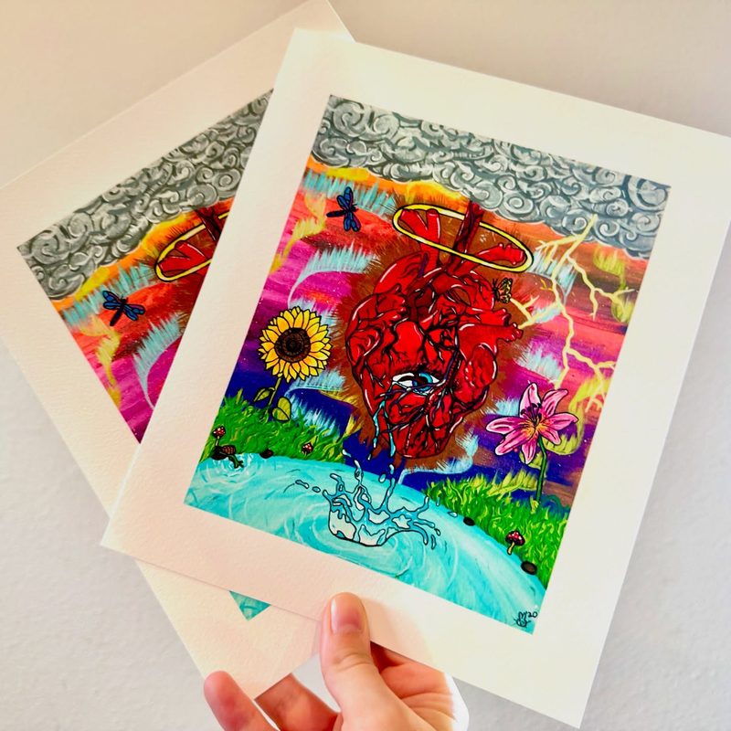 A hand holding two giclée prints of The Biggest Hearts Carry the Heaviest Burdens with white paper borders.