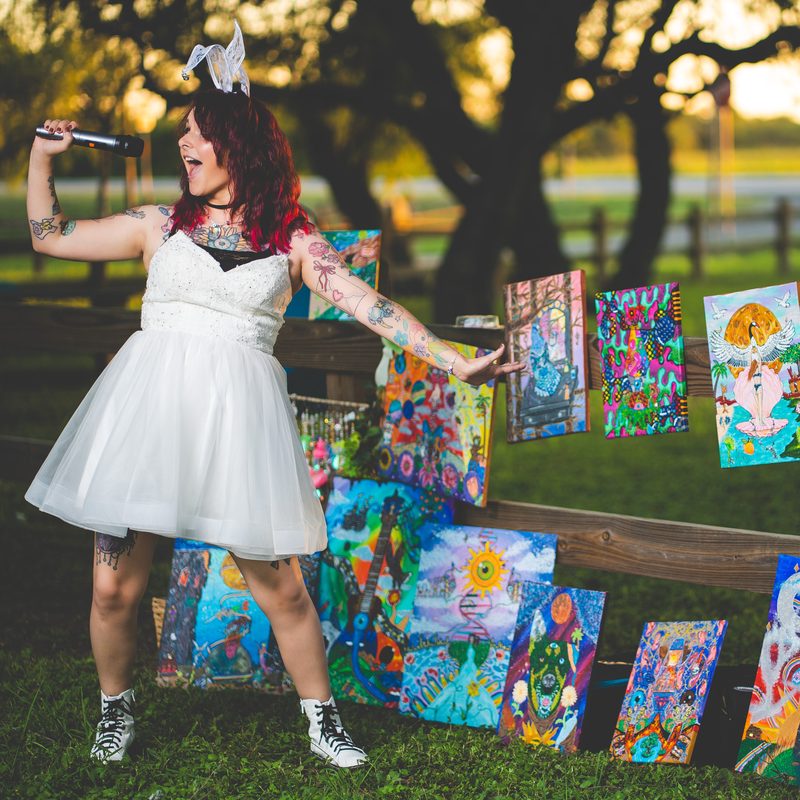 Shai in a white dress and bunny ears, singing into a microphone beside a fence lined with her paintings