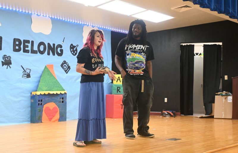Shai on a school stage in front of an I Belong mural, speaking beside poet Kiya Vance